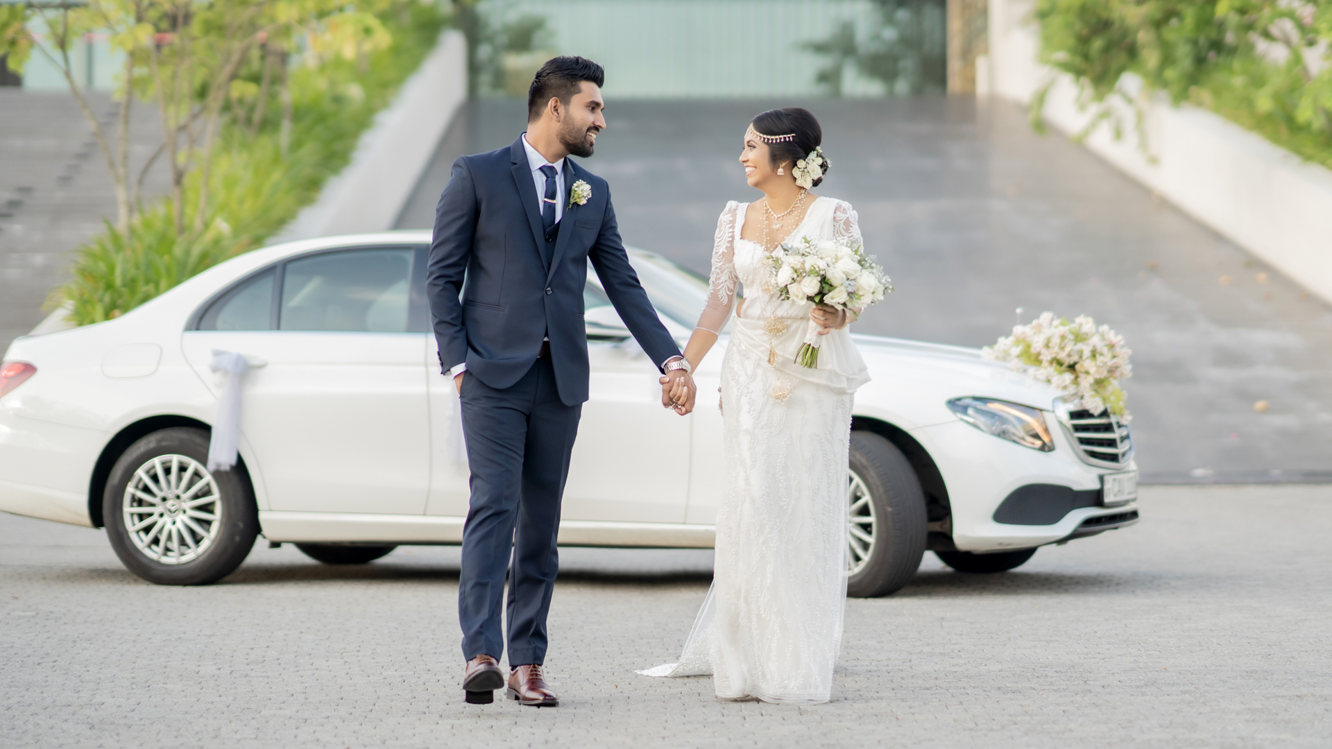 A bride and groom in beautiful wedding outfits, holding hands and smiling, standing in front of a decorated white car on their wedding day, mangala yojana 2024, matrimony sri lanka, sl marriage proposals, mangala yojana, marriage proposal in sri lanka, sri lankan matrimony, sri lankan marriage proposal sites