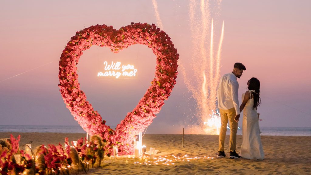 Romantic beach proposal with a heart-shaped flower arch and "Will you marry me?" sign, fireworks in the background, and a couple holding hands.
