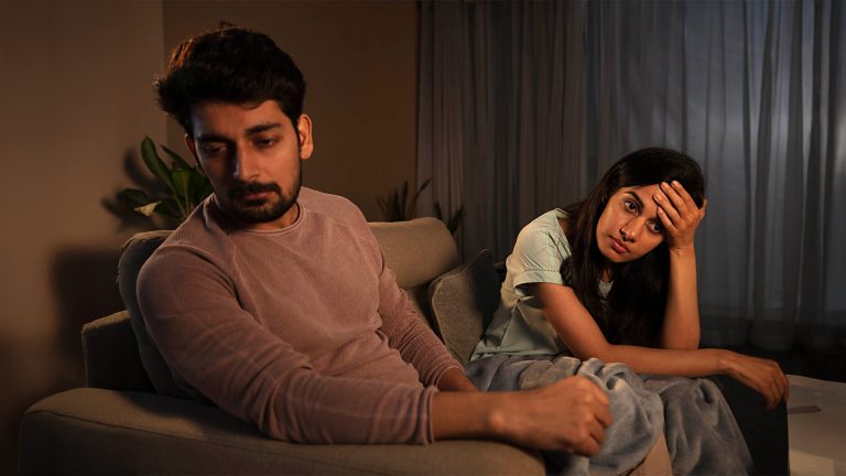 A man and woman sit on a couch looking upset and distant, with the woman holding her head and the man looking away.
