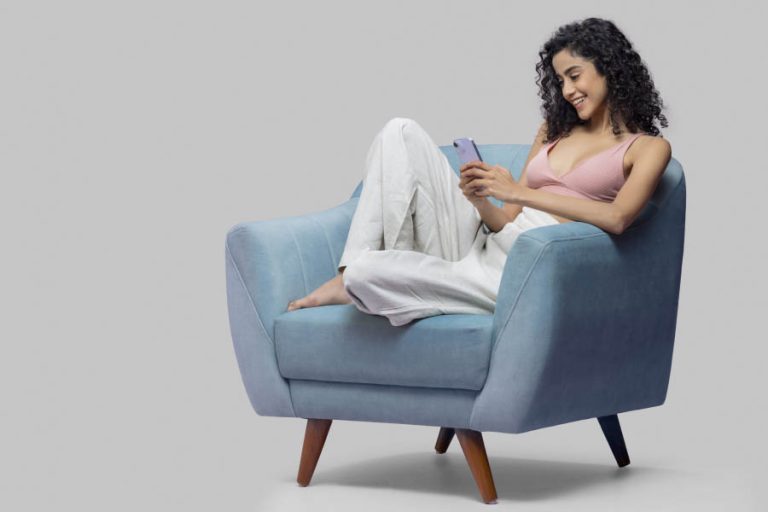 A woman sitting in a blue chair, focused on her phone with a relaxed expression, browsing marriage proposals online.